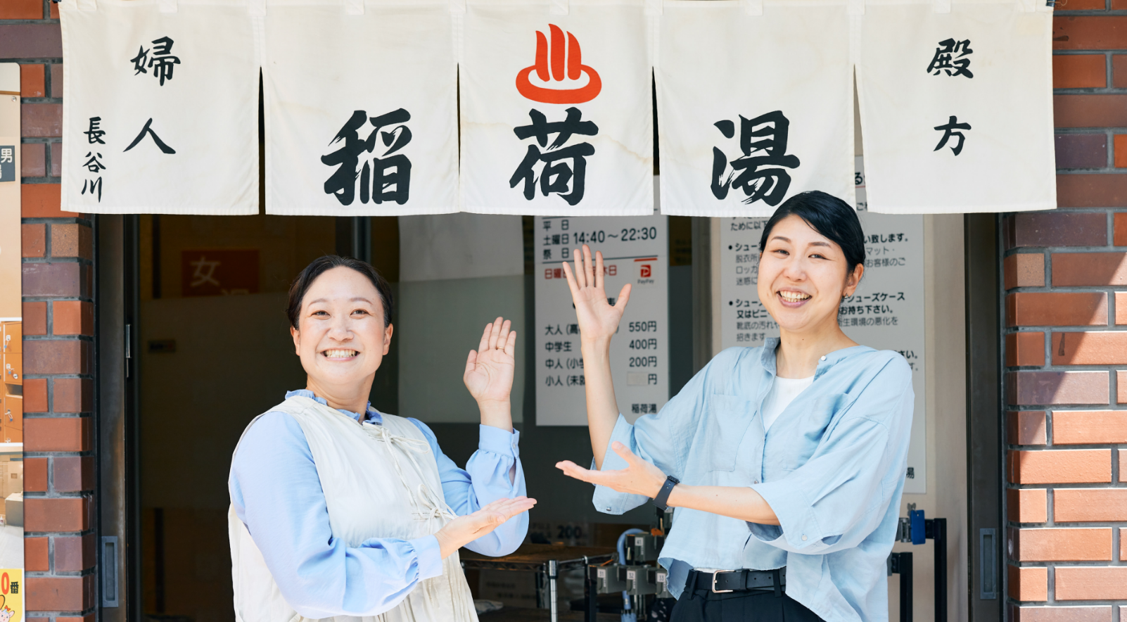 稲荷湯の暖簾の前でポーズをとる川島さんと田中さん